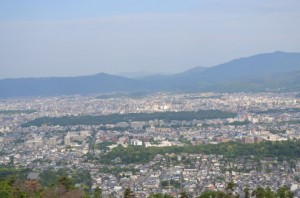 A panoramic view of a city with numerous buildings and houses, surrounded by greenery and trees. In the background, there are mountain ranges under a clear sky.