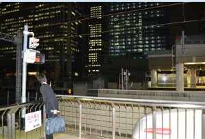 A person in a suit is standing on a platform at a train station at night, holding a blue bag. The background features tall buildings with many illuminated windows, indicating an urban setting. There is a sign in Japanese on the railing near the person.