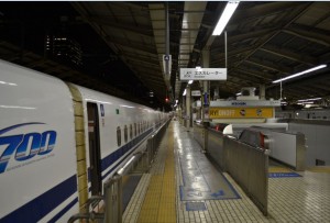 The image shows a train station platform at night. A white high-speed train, marked with "700" on its side, is stationed on the left side of the platform. The platform is well-lit with overhead lights and has tiled flooring. There are signs hanging from the ceiling, and another train is visible on the right side of the platform. The station appears to be clean and orderly, with safety barriers and markings on the ground.