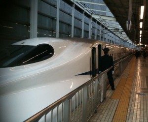 The image shows a sleek, white high-speed train at a station platform. A person in a uniform, possibly a train conductor or staff member, is standing near the open door of the train. The platform is tiled and has a yellow tactile paving strip along the edge. The station is covered and well-lit, with other people visible in the background.