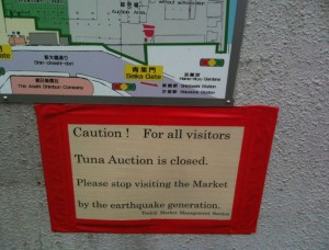 The image shows a sign with a map above it. The sign reads: "Caution! For all visitors Tuna Auction is closed. Please stop visiting the Market by the earthquake generation. Tsukiji Market Management Section." The sign is bordered in red and is posted on a gray wall. The map above the sign appears to be a layout of the market area.