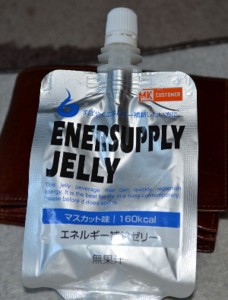 The image shows a silver pouch of "Enersupply Jelly," a consumable energy gel product. The pouch has a spout at the top for easy consumption. The packaging includes text in both English and Japanese, indicating that the product is muscat-flavored and contains 160 kcal. The background includes a brown leather item, possibly a wallet or a case.