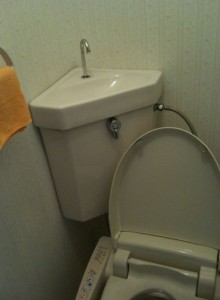 The image shows a toilet with an integrated sink on top of the toilet tank. The sink faucet is positioned above the tank, allowing water to flow into the tank after use. There is also a yellow towel hanging on a rack on the wall to the left of the toilet. The toilet seat is open, and the setup appears to be in a small bathroom.