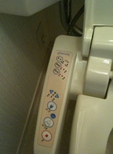 The image shows the control panel of a Japanese-style bidet toilet seat. The panel has various buttons with icons and text, likely indicating different functions such as water spray, bidet, and drying options. The buttons are arranged vertically and include both symbols and Braille. The toilet seat is white, and the control panel is attached to its side.