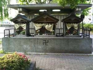 The image shows a small shrine with three wooden structures, each adorned with white curtains and floral arrangements. The structures are elevated on a concrete platform, and there is a swastika symbol on the front of the platform. The shrine is situated outdoors, surrounded by greenery and a paved area.