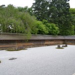 This image shows a traditional Japanese rock garden, also known as a Zen garden. The garden features a flat expanse of white gravel, meticulously raked to create patterns. Several large rocks are strategically placed throughout the gravel, some surrounded by small patches of moss. In the background, there is a wooden wall with a natural, weathered appearance, and behind the wall, lush green trees and foliage are visible. The overall scene is serene and orderly, typical of Zen garden aesthetics.