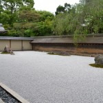 The image shows a traditional Japanese rock garden, also known as a Zen garden. The garden features a flat expanse of white gravel, meticulously raked into patterns. There are several clusters of rocks and small patches of moss strategically placed within the gravel. The garden is enclosed by a wooden wall, and there is a traditional Japanese building with a tiled roof on the left side. In the background, there are lush green trees and foliage. The overall scene is serene and minimalist, typical of Zen gardens designed for meditation and contemplation.