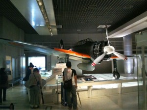 The image shows a museum exhibit featuring a vintage aircraft. The aircraft is displayed indoors, with several people standing around it, observing and taking pictures. The plane has a propeller and is painted with a combination of black, white, and orange colors. The museum setting includes informational displays and a well-lit environment.