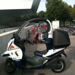 A covered scooter is parked in an outdoor area next to a concrete wall. The scooter has a roof and a windshield, and it is branded with the BMW logo and the model name "C1 200." In the background, there are several bicycles parked in a bike rack, and a red scooter is also visible. Trees and a modern building are in the distance.