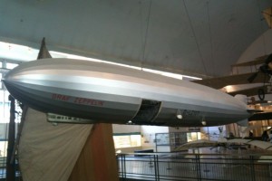 The image shows a model of a zeppelin airship displayed in a museum. The zeppelin is silver in color and is suspended from the ceiling. The words "Graf Zeppelin" are visible on the side of the airship. The background includes other exhibits and museum displays.