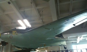 The image shows a vintage military aircraft displayed indoors, possibly in a museum. The aircraft has a sleek, streamlined design with a single wing extending from the fuselage. The lighting in the room is bright, with fluorescent lights visible on the ceiling. The aircraft appears to be suspended or mounted for display purposes.