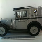 Alt text: A vintage BMW delivery van from 1930, labeled "BMW 3/15 PS," is displayed in a museum. The van is gray with black fenders and features the BMW logo and the text "Teile in Eile" and "BMW AG München" on its side. The vehicle has a boxy shape with a roof rack and large round headlights.