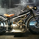 The image shows a vintage motorcycle with a black and white color scheme. It features a brown leather seat and a prominent engine with visible mechanical components. The motorcycle is displayed against a backdrop that appears to be an old workshop or industrial setting, with various tools and materials in the background. The overall appearance suggests that the motorcycle is a classic or antique model, likely from the early to mid-20th century.