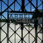 Alt text: A view through the iron gate of the Dachau concentration camp memorial site, with the infamous phrase "Arbeit Macht Frei" (Work Sets You Free) inscribed on the gate. In the background, visitors are walking around the open grounds and buildings of the memorial site under a clear blue sky.