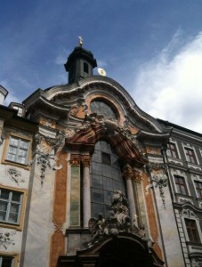 The image shows the ornate facade of a historic building with baroque architectural elements. The building features intricate carvings, statues, and decorative details around a large arched window. The facade is adorned with various colors and textures, including marble-like columns and painted surfaces. A small tower with a clock and a golden statue is visible at the top of the building. The sky above is partly cloudy.