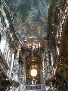 The image shows the interior of a highly ornate baroque church. The ceiling is elaborately painted with religious scenes and figures, and the walls are adorned with intricate carvings, gilded decorations, and statues. A large, circular stained glass window is visible in the center, allowing light to illuminate the richly decorated space. The overall atmosphere is grand and opulent, characteristic of baroque architecture.