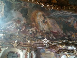 The image shows an ornate ceiling with intricate frescoes and detailed artwork. The ceiling is richly decorated with gold accents and features various figures and scenes, likely of religious or historical significance. The architecture includes arched windows and elaborate moldings, contributing to the grandeur of the space. The overall atmosphere is one of opulence and artistic mastery.