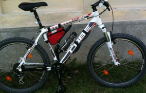 The image shows a white and black mountain bike with red accents, leaning against a wall. The bike has a water bottle mounted on the frame and a small red bag attached to the top tube. The wheels have orange reflectors. The bike is positioned on a grassy area.