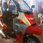 The image shows a red and silver BMW C1 scooter. The scooter has a unique design with a roof and a windshield, providing some protection from the elements. It features a single front wheel and a single rear wheel, with a seat and handlebars for the rider. The BMW logo is visible on the side of the scooter. The background appears to be a display or showroom setting.