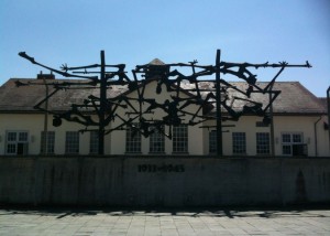 The image shows a large, abstract metal sculpture mounted on a concrete wall in front of a building. The sculpture consists of numerous intertwined and elongated human-like figures, creating a chaotic and somber scene. Below the sculpture, the years "1933-1945" are inscribed on the wall. The building behind the sculpture has a pitched roof and several windows. The sky above is clear and blue.