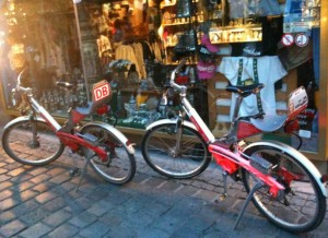 The image shows three red and white bicycles parked on a cobblestone street in front of a shop window. The shop window displays various items, including clothing and accessories. The bicycles have a sign with the letters "DB" on them, indicating they might be part of a bike-sharing or rental program. The scene appears to be in an urban area.