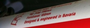 The image shows a close-up of a white and red object with red text printed on it. The text reads: "Alp Rider 430mm, Fronttravel 100mm, Designed & engineered in Bavaria." There is also a small graphic of a bicycle frame next to the text.