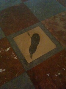 The image shows a tiled floor with a pattern of alternating dark and light squares. One of the light-colored tiles in the center has a dark, irregularly shaped stain or mark on it, resembling a footprint or a smudge. The surrounding tiles are a mix of reddish-brown and dark gray colors.