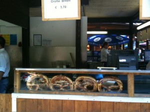 The image shows a food stall with a display case containing large pretzels. There is a sign above the display case that reads "Große Brezn € 3,70." In the background, there are people working behind the counter, and a blue umbrella with white text is visible. The stall appears to be part of an outdoor market or festival setting.