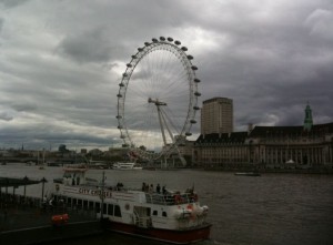 The image shows a large Ferris wheel, known as the London Eye, situated on the bank of the River Thames in London. In the foreground, there is a riverboat named "City Cruises" docked at the riverbank. The sky is overcast with dark clouds, and several buildings are visible in the background.