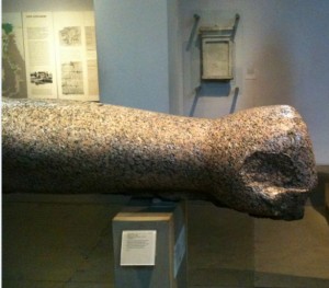 The image shows a large, ancient stone artifact that appears to be a broken statue or sculpture, displayed in a museum setting. The artifact is mounted on a pedestal with an informational plaque. The background includes informational displays and a wall-mounted exhibit. The stone has a rough, weathered texture and is partially broken off at one end.