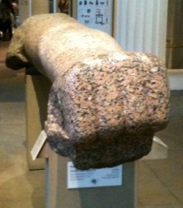 The image shows a large, ancient stone artifact displayed in a museum. The artifact appears to be a broken or eroded statue, possibly of a human or animal figure, made from a speckled, reddish-brown stone. It is mounted on a pedestal with informational plaques attached to it. The background includes other museum displays and a column.