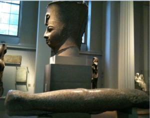 The image shows a museum exhibit featuring a large stone bust of an ancient Egyptian pharaoh, prominently displayed on a pedestal. In the foreground, there is a large stone arm, likely part of a statue, also on display. The background includes other artifacts and sculptures, with a window providing natural light to the room.