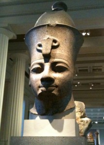 The image shows a large stone bust of an ancient Egyptian pharaoh, displayed in a museum setting. The pharaoh is wearing a traditional headdress with a cobra emblem on the front. The background includes tall columns and other museum exhibits.