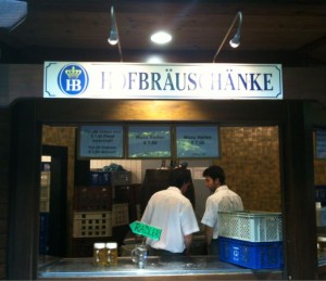 The image shows a food or drink counter with a sign above it that reads "Hofbräuschänke." Two people in white shirts are working behind the counter. There are crates and containers around them, and two glasses of beer are placed on the counter. The setting appears to be a casual dining or bar area.