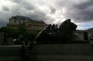 A group of people is sitting on a large lion statue in an urban area. The sky is cloudy, and there are buildings in the background. The statue appears to be part of a larger monument or public square.