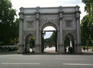 The image shows the Marble Arch, a white marble monument with three arches, located in London. The structure features intricate carvings and is set against a backdrop of trees and a partly cloudy sky. There are people visible near the arch, and a road with white lane markings runs in front of it.