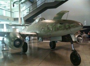 The image shows a military aircraft displayed indoors, likely in a museum. The aircraft has a sleek, metallic body with a camouflage pattern and is equipped with two jet engines mounted under its wings. The landing gear is extended, and there is a staircase and railing in the background, indicating an indoor exhibit space.