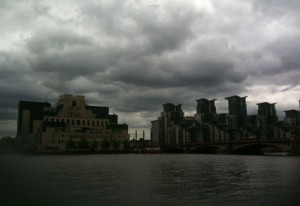 The image shows a waterfront cityscape under a cloudy sky. On the left side, there is a large, distinctive building with a unique architectural design, featuring multiple levels and a blocky structure. To the right, there are several modern high-rise buildings with a uniform appearance, characterized by their tall, narrow shapes and flat roofs. The water in the foreground reflects the overcast sky and the buildings.