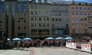 The image shows a crowd of people gathered in front of a row of multi-story buildings. The buildings have various architectural styles and are closely packed together. There are several blue umbrellas providing shade for the people, and a white and red barricade is visible on the right side of the image. The scene appears to be in an urban area, possibly a city square or a pedestrian zone.