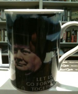 A white ceramic mug with a black-and-white image of a man's face on it. Below the image, there is text that reads, "LET US GO FORWARD TOGETHER." The mug is placed on a white surface, and there are bookshelves with books in the background.