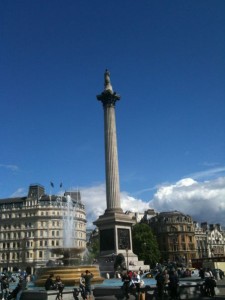 The image shows a tall column with a statue on top, located in a public square. There are fountains and people gathered around the base of the column. Surrounding the square are several historic buildings with classic architecture. The sky is clear with a few clouds.