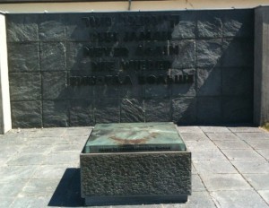 The image shows a memorial with a stone wall in the background and a rectangular stone pedestal in the foreground. The wall has an inscription that reads, "THEY FOUGHT FOR JAPAN NEVER AGAIN WILL THERE BE A HIROSHIMA BOMB." The pedestal also has an inscription, but it is not fully visible in the image. The area is paved with stone tiles.