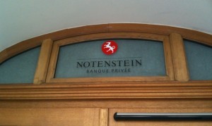 The image shows a wooden-framed glass panel above a door. The glass panel has the text "NOTENSTEIN BANQUE PRIVÉE" written on it, with a red circular logo featuring a white lion above the text.