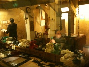 The image shows a room with a vintage or historical setting, likely a wartime operations room or command center. There are several old-fashioned telephones of various colors on a long desk, along with papers and other office supplies. Two people are present: one person is seated at the desk, appearing to be using a telephone, while the other person is standing in the background, looking at a wall with maps or documents pinned to it. The room has a cluttered and busy atmosphere, with dim lighting and a utilitarian design.