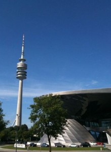 The image shows a tall communications tower with a distinctive design, standing next to a modern, curved building. There are trees and parked cars in the foreground, and the sky is clear and blue.