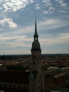 The image shows a tall church tower with a green spire and clock faces on its sides, set against a backdrop of a cityscape with numerous buildings and a partly cloudy sky. The city extends into the distance, with rooftops and other structures visible under the blue sky with scattered clouds.