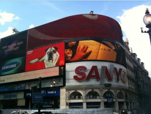 The image shows a large building with multiple electronic billboards displaying various advertisements. The prominent ads include a Sanyo sign, a Samsung ad, and a McDonald's ad featuring a dog and a hand reaching for food. The sky is partly cloudy, and there is a street lamp visible on the right side of the image. The building has an arched architectural design on the lower level.