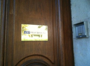 A wooden door with a brass plaque that reads "PKB PRIVATBANK AG" is shown. To the right of the door, there is an intercom system mounted on a concrete wall.