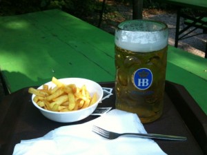 A large glass of beer with a frothy head sits on a tray next to a bowl of French fries. A fork and a napkin are also on the tray. The background features green picnic tables and a shaded outdoor setting.