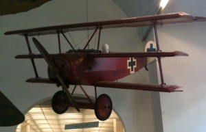 A red triplane aircraft is displayed hanging from the ceiling in a museum. The plane has three wings stacked vertically and features a black cross insignia on its side. The background shows part of the museum's interior with an arched doorway and some lighting.